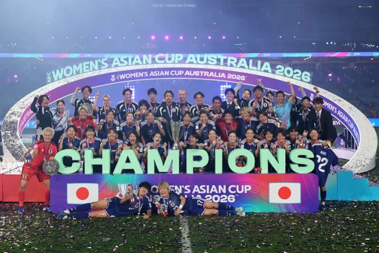 Japanese women’s national football team players in blue jerseys cheering and lifting the AFC Women’s Asian Cup trophy on a podium amidst gold confetti at Stadium Australia.