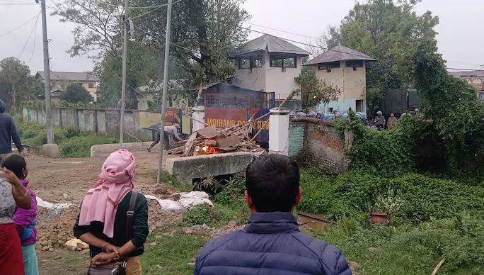 Protest outside the damaged Moirang Police Station, Manipur, on April 7, 2026. A fire burns at the gate as police in riot gear watch from within.