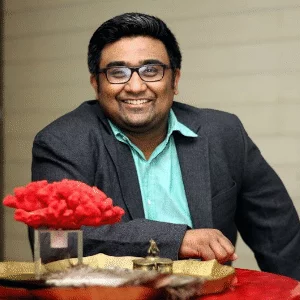 An Indian entrepreneur, Kunal Shah, wearing glasses, a teal button-up shirt, and a dark grey blazer, smiling while leaning forward slightly at a table.
