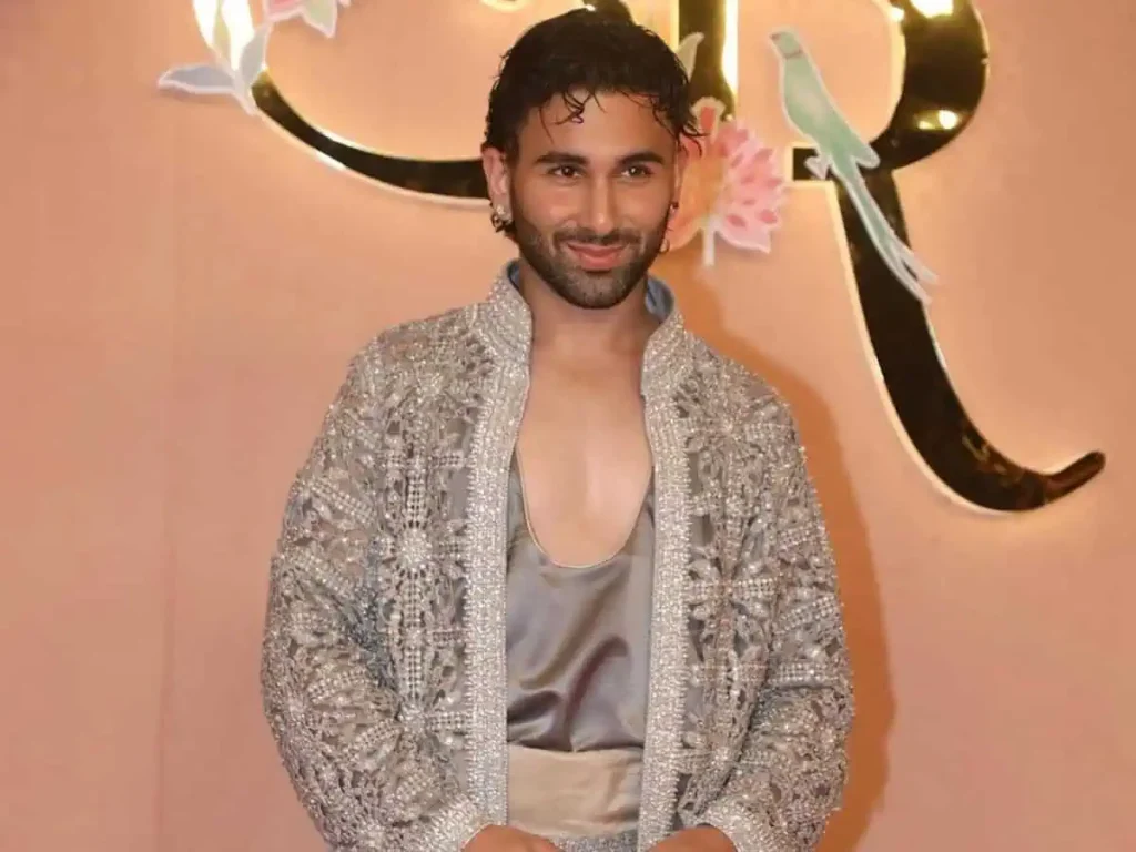A medium shot of Orry smiling and posing against a peach-colored backdrop. He is wearing a heavily embellished silver-grey sheer jacket over a low-cut satin top.