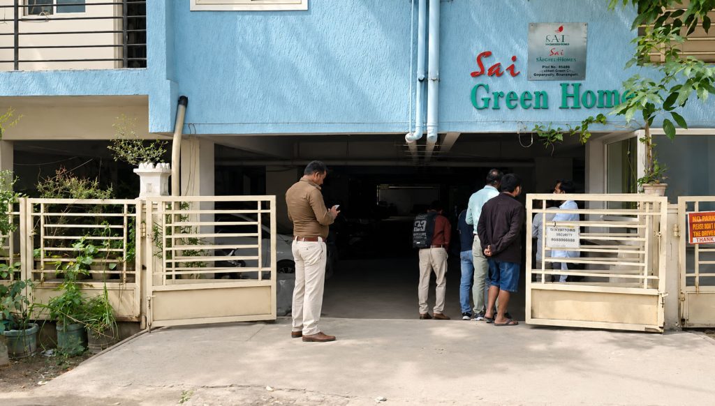 Police personnel and onlookers gathered outside the open gated entrance of the "Sai Green Homes" apartment building.