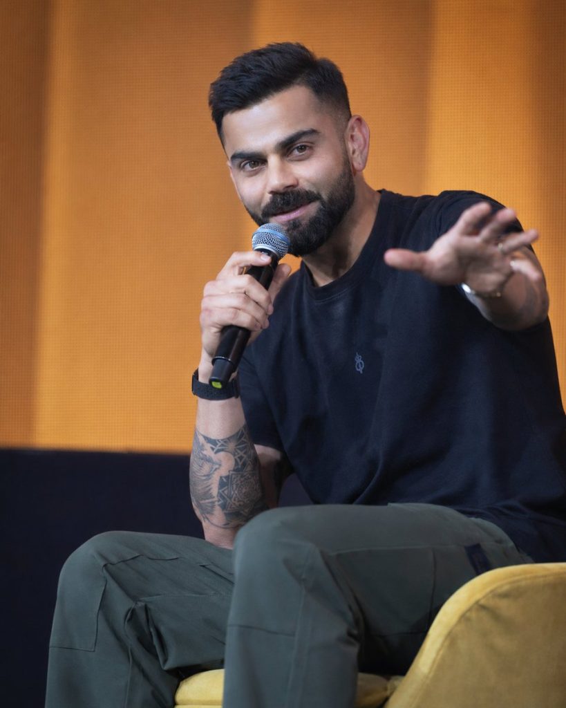 Virat Kohli sitting on a yellow chair in a black t-shirt and green trousers, holding a microphone in his right hand and gesturing with his left hand against an orange background.
